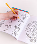 Model holds a yellow-colored pencil above a graphic of various breakfast items on a page of the Pusheen coloring book. The book is open to show the lefthand page graphic and part of the righthand page. On the lefthand page showcases Pusheen as eight different breakfast items with character descriptors under each. On the righthand page is a circular graphic where Pusheen takes the form of an avocado toast, bagel, cereal, and sunny-side-up egg accompanied by forks, spoons, strawberries, and sparkles. 