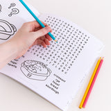 Model holds a blue colored pencil above a crossword puzzle page of the Pusheen activity book. The crossword puzzle has many bread-themed words including croissant. Sourdough, and toast. Under the crossword puzzle is a graphic of a breadbasket holding Pusheen taking the form of a baguette, croissant, and loaf of bread.  