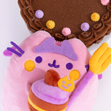 Pusheen Devil's Food Cake Plush lays on a white surface. The light pink and purple plush is holding a fork and cake in her front paws. The crowned plush is surrounded by a chocolate cake decorated with pink, purple, and orange icing dollops. 