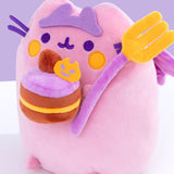 Close-up view of felt crown and the plush items Pusheen is holding. The brown and yellow layered cake plush is topped with purple icing, a chocolate frosting dollop, and a felt orange pumpkin. The large plush fork has a purple handle with yellow tines. 