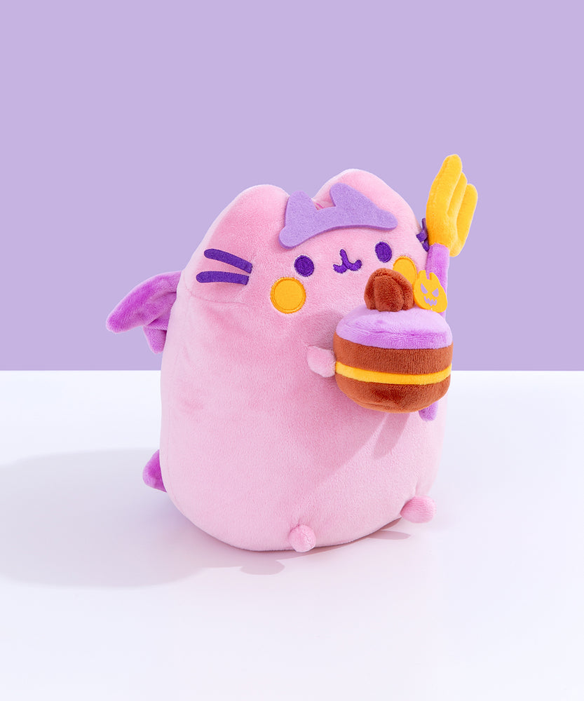 Right quarter view of the Devil’s Food Cake Plush showing Pusheen the Cat holding a plush layered cake in her front paws. The cake is topped with a felt pumpkin and brown icing dollop. From this view, Pusheen’s wings and tail can be seen coming out of her back. 