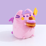 Right quarter view of the Devil’s Food Cake Plush showing Pusheen the Cat holding a plush layered cake in her front paws. The cake is topped with a felt pumpkin and brown icing dollop. From this view, Pusheen’s wings and tail can be seen coming out of her back. 