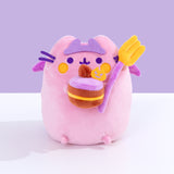 Devil’s Food Cake Pusheen Plush sits atop a white surface in front of a purple background. The pink plush holds a yellow and purple fork and a layered devil’s food cake. 