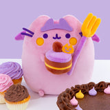 Front view of Devil’s Food Cake Pusheen Plush. The plush a light pink and purple Pusheen smiling. The cat plush wears a light purple crown while holding a purple and yellow fork and a yellow, brown, and purple layered cake. Pusheen’s purple facial features and paws extend off her body. The Plush sits on a white surface surrounded by purple, orange, and pink sweet treats.