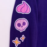 Close-up view of right sleeve of the Halloween Pusheen long-sleeve tee. The graphic running down the sleeve features three icons: a pink whipped frosting dollop, a purple and pink skull, and yellow sparkles that are all outlined in purple and white.  