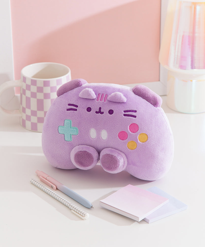 Gaming controller plush sits on a desktop. The purple plush has darker purple trigger buttons on top of the plush and joysticks near the bottom of the plush. 