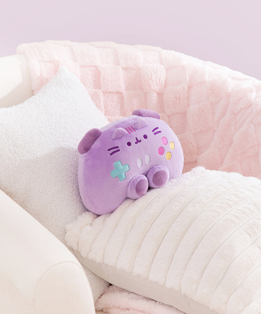 Gamer Pusheen Controller Plush sits in a chair surrounded by blankets and pillows to show the size and scale of the purple plush toy.