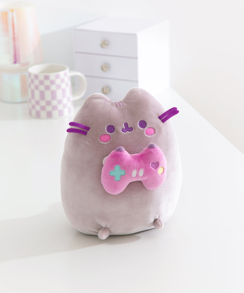 Gamer themed cat plush sits on a white desktop. The purple-hued cat holds a pink gaming controller with multi-colored buttons.