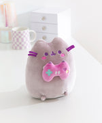 Gamer themed cat plush sits on a white desktop. The purple-hued cat holds a pink gaming controller with multi-colored buttons.