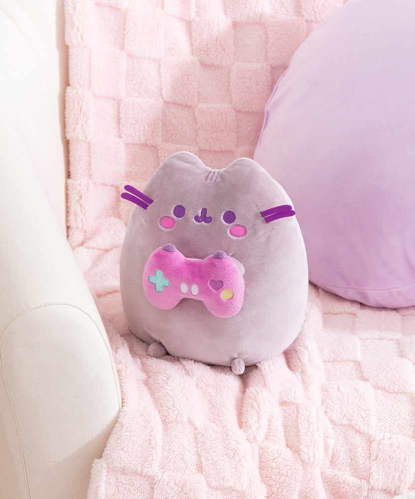Alternate side quarter view of gamer plush. Pusheen the Cat holds a pink and purple gaming controller with a teal plus button, purple heart button, yellow circle button, and two white oblong buttons on the front of the controller.