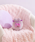 Gamer Pusheen Plush sits in a white chair with a purple pillow and pink blanket behind the plush to show the size and scale of the gaming plush.