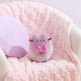 Gamer Pusheen Plush sits in a white chair with a purple pillow and pink blanket behind the plush to show the size and scale of the gaming plush.