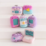 Seven gaming themed mini plush keychains sit atop a white pedestal. The assortment includes Pusheen the Cat taking the form of an accessory case, a bag of chips, two different handheld gaming consoles, and energy drink, a controller, and a laptop.