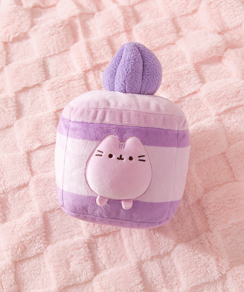 Purple cake plush lies on a pink textured surface. Pusheen transforms into a layered lavender-flavored mini cake. 