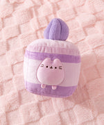 Purple cake plush lies on a pink textured surface. Pusheen transforms into a layered lavender-flavored mini cake. 
