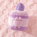 Purple cake plush lies on a pink textured surface. Pusheen transforms into a layered lavender-flavored mini cake. 
