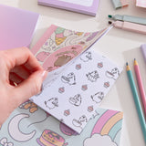 Model holds open the Pusheen activity book showcasing one of the coloring pages. The open page shows a black outline Pusheen carrying cupcakes, mixing batter, and mini cupcakes.