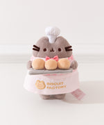Pink Biscuit Baker Pusheen Plush in front of a white background. Pusheen holds her three biscuit buns on a gray platter. Pusheen’s body is a fuzzy grey material. The Pusheen corp tag can be seen coming off the bottom right of the plush.