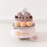 Pink Biscuit Baker Pusheen Plush in front of a white background. Pusheen holds her three biscuit buns on a gray platter. Pusheen’s body is a fuzzy grey material. The Pusheen corp tag can be seen coming off the bottom right of the plush.