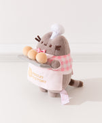 Side view of the Pusheen plush. Pusheen's front paws are holding a tray of biscuits that extends off of the front of the standing plush. 