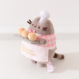 Side view of the Pusheen plush. Pusheen's front paws are holding a tray of biscuits that extends off of the front of the standing plush. 