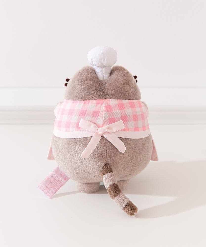 Back view of Pink Baker Pusheen Plush. The checkered top and apron combo are tied in the back with a bow. Pusheen’s gray striped tail is seen coming off the bottom back of the plush. 