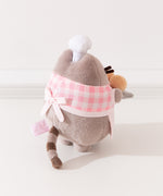 Alternate back quarter view of plush. Pusheen wearing a baking apron tied with a bow in the back while the plush stands upright on two pudgy paws. 