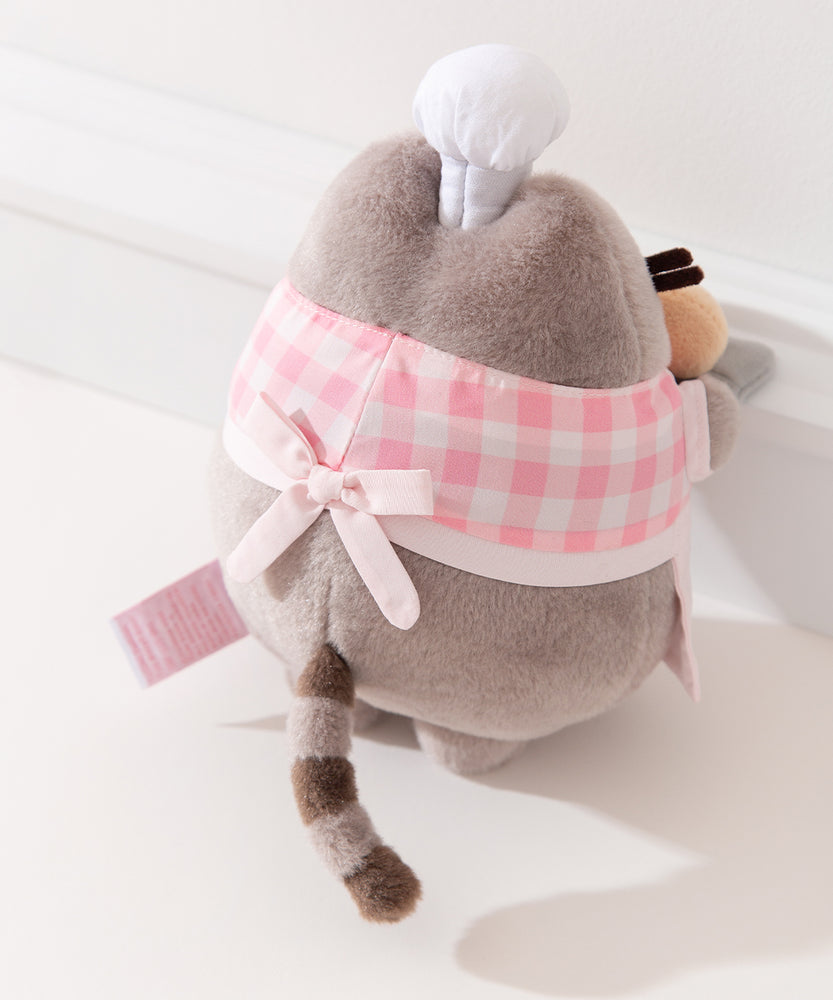 Back view of the Pink Biscuit Baker Pusheen Plush. The checkered top and apron combo are tied in the back with a bow. Pusheen’s gray striped tail is seen coming off the bottom back of the plush. 