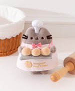 Pusheen Biscuit Baker Plush has a light grey fuzzy texture to mimic the Cat's fur. The standing plush features a mini white chef's hat, pink gingham checkered shirt with white lapels, mini pink bowtie, and white apron that says "Biscuit Factory."