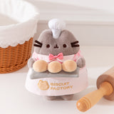 Pusheen Biscuit Baker Plush has a light grey fuzzy texture to mimic the Cat's fur. The standing plush features a mini white chef's hat, pink gingham checkered shirt with white lapels, mini pink bowtie, and white apron that says "Biscuit Factory."