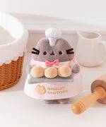 Pink Biscuit Baker Pusheen Plush in front of a white background. Pusheen the Cat shows off her biscuit baking skills while wearing a pink and white gingham shirt with an pink bowtie and an apron that says “Biscuit Factory.”