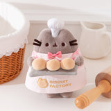 Pink Biscuit Baker Pusheen Plush in front of a white background. Pusheen the Cat shows off her biscuit baking skills while wearing a pink and white gingham shirt with an pink bowtie and an apron that says “Biscuit Factory.”