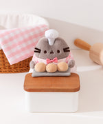 Close-up of Pusheen holds three biscuit buns on a grey slab on a wood countertop. Pusheen wears a white patisserie chef’s hat between her two fuzzy gray ears. 