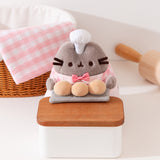 Close-up of Pusheen holds three biscuit buns on a grey slab on a wood countertop. Pusheen wears a white patisserie chef’s hat between her two fuzzy gray ears. 