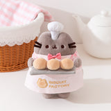 Front view of Pink Biscuit Baker Pusheen Plush. The grey fluffy Pusheen stands in front of a white tea pot, whicker bread basket, and a pink gingham hand towel.