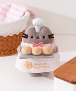 Biscuit Baker Pusheen Plush in front of a wears a white apron with a gold embroidered logo of "Biscuit Factory" with an outline of a cat's head next to the text.