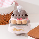 Biscuit Baker Pusheen Plush in front of a wears a white apron with a gold embroidered logo of "Biscuit Factory" with an outline of a cat's head next to the text.