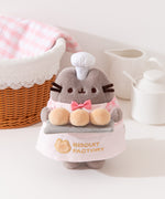 A cat plush wears a tiny pink bowtie and white chef's hat.