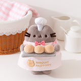 A cat plush wears a tiny pink bowtie and white chef's hat.