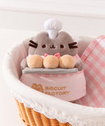 Pink Biscuit Baker Plush sits in a bread basket. The golden yellowcolor of the biscuits Pusheen holds matches the embroidery color of the Biscuit Factory logo.
