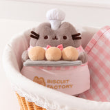 Pink Biscuit Baker Plush sits in a bread basket. The golden yellowcolor of the biscuits Pusheen holds matches the embroidery color of the Biscuit Factory logo.