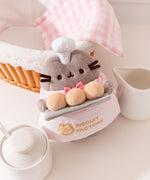 Top aerial view showcasing Pusheen’s face, fuzzy coat texture, and her white patisserie chef hat that sits between her triangular ears. 