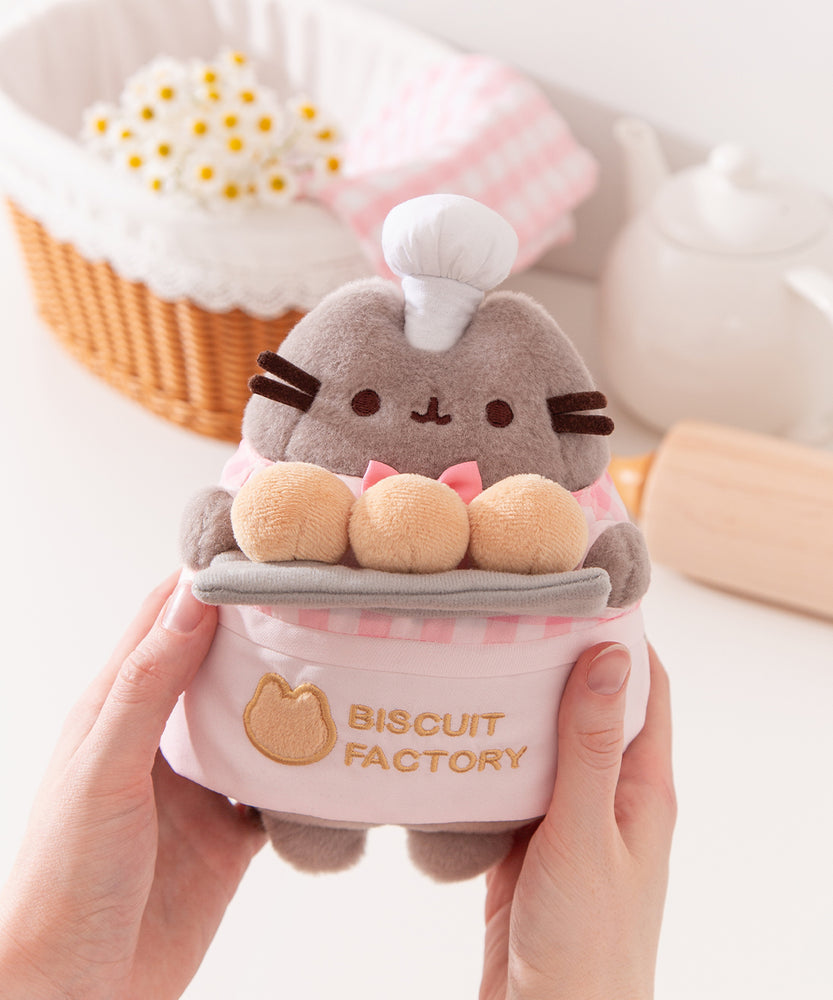 Model holding Biscuit Baker Pusheen Plush to show size scale. The sides of the plush fit in the model’s hands while the head and legs are not encompassed by the hands. 