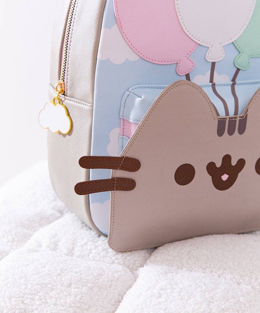 Side and back view of the backpack. The pack has gray adjustable straps with golden hardware. The back of the pack features a graphic of Pusheen the Cat floating through a cloudy sky. 