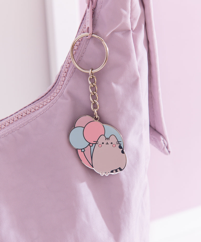 Flat ring keychain depicting Pusheen the cat with three pastel balloons hangs off a purple purse. 