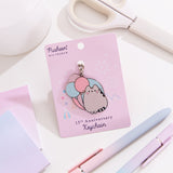 Pusheen 15th anniversary shiny nickel keychain attached to its light purple backer card. The background shown on the paper card is of a festival scene with fair rides.