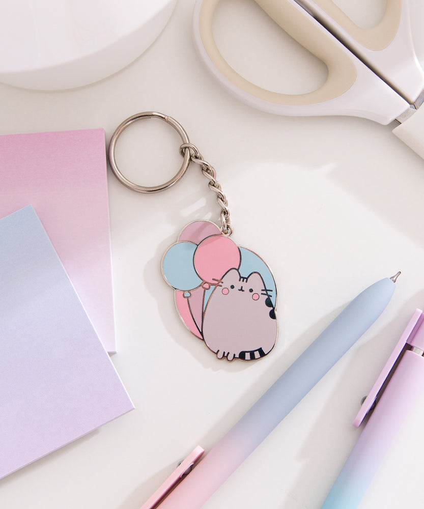 Pusheen keychain charm with flat split ring attached to a chain and decorative Pusheen plate. A blushing grey cat sits in front of pink, purple, and blue balloons. 