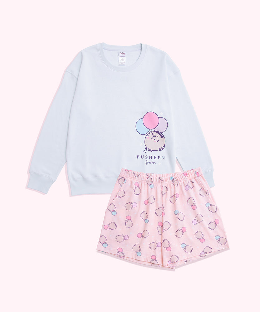 Pusheen 15th Anniversary Pajama Set lies as a set on a background. The oversized light blue sweatshirt has a graphic of Pusheen the Cat with balloons on the wearer's left. The stretchy patterned shorts have an all over pattern of Pusheen holding pastel balloons. 