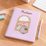 Front cover of the Pusheen 16-month planner on a wooden desk with stationery items. The pink planner shows a graphic of Pusheen looking in a mirror with the phrase "you look good".