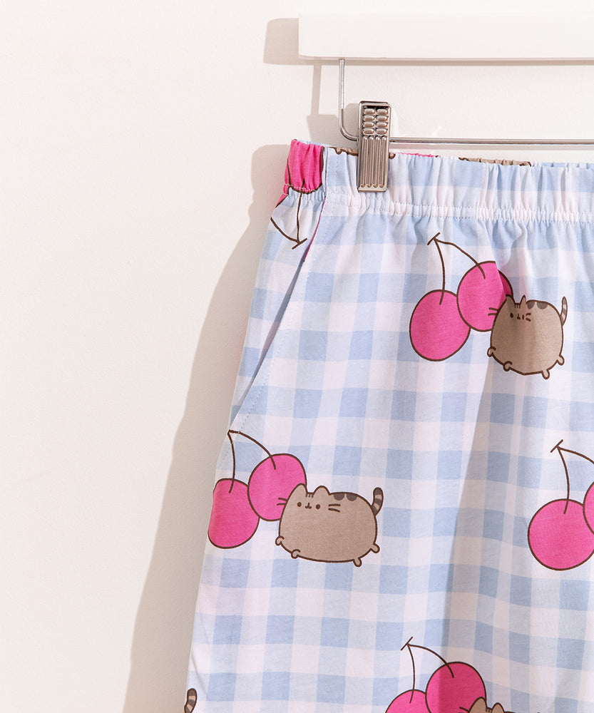 Pusheen Cherry Pajama Set includes a white and blue gingham oversized top and elastic waistband shorts. The set has an all over print of Pusheen the Cat leaping next to two dark pink cherries. 